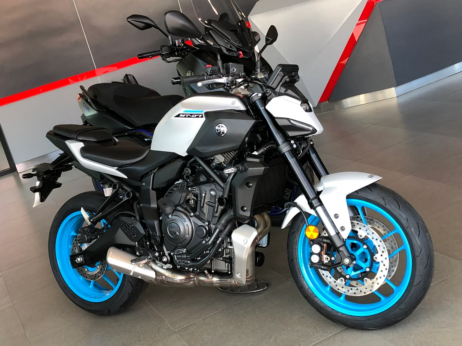 White and cyan 2025 Yamaha MT-07 Y-AMT photographed in a showroom, viewed from the front right.